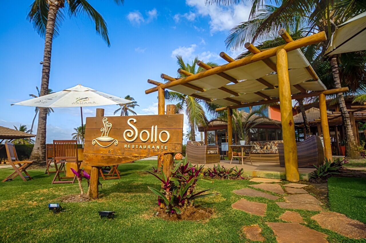 Señal de madera que indica 'Sollo Restaurante', con un entorno de jardín tropical, palmeras y mesas al aire libre.