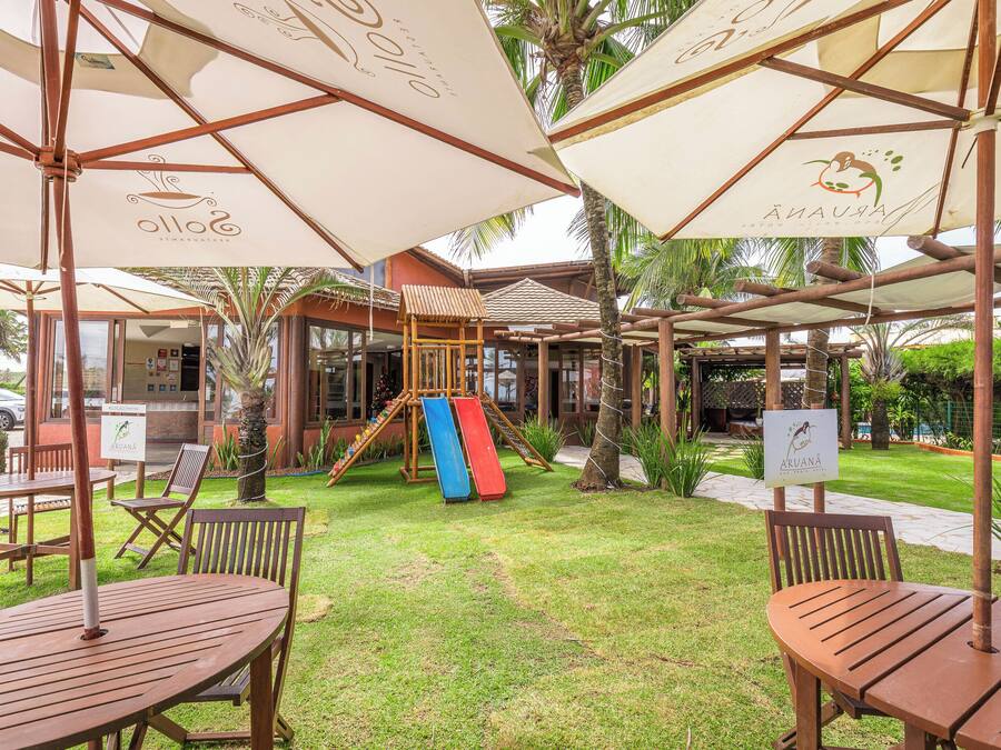 An outdoor seating area with wooden tables and chairs, umbrellas, and a children's play structure.