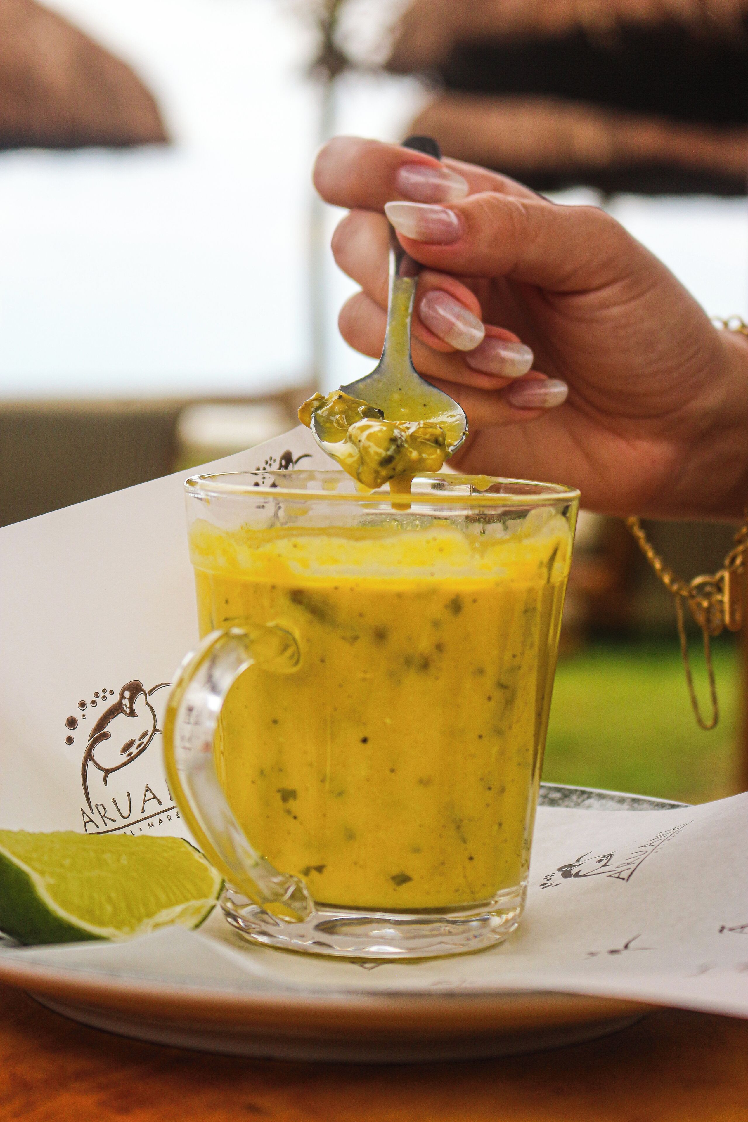 Una mano sosteniendo una cuchara sobre un vaso de batido amarillo con una cuña de lima al lado.