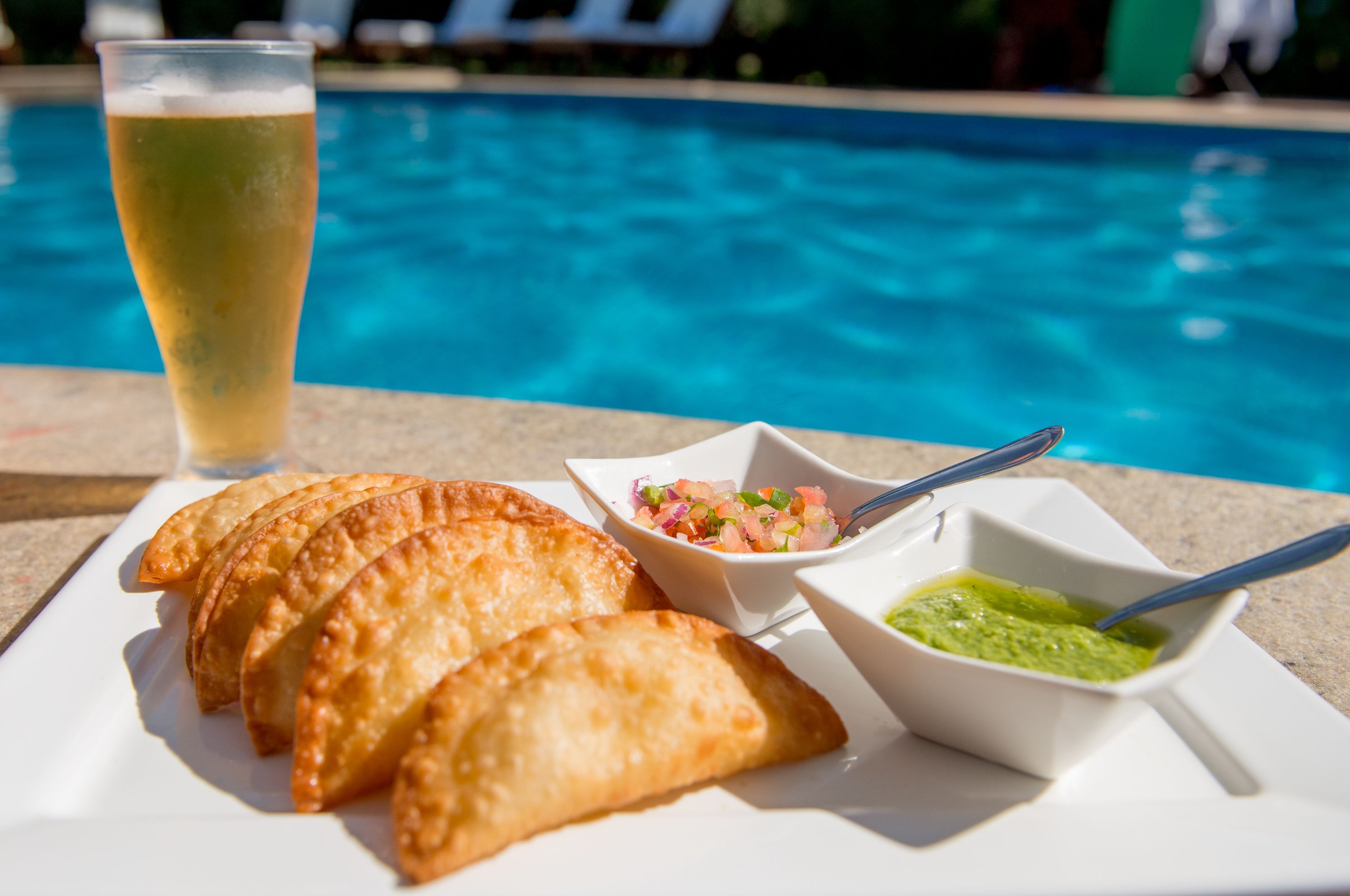 A plate of fried pastries, a glass of beer, and two small bowls of dips by a poolside.