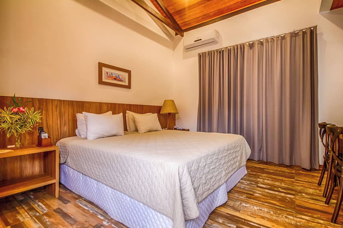 A hotel room with a large bed, wooden headboard, a bedside table with a lamp, a picture on the wall, and a window with curtains.