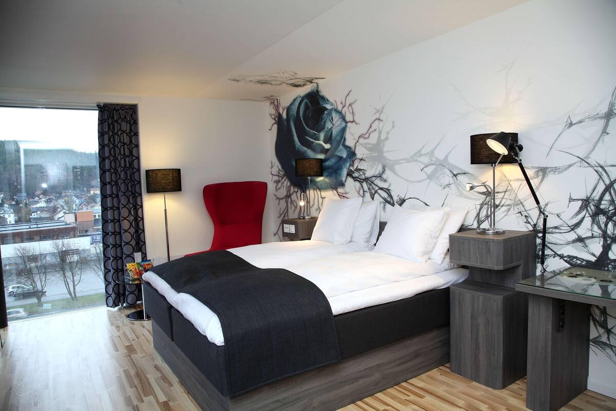 A modern bedroom with a large bed, a red chair, and a city view through a window.