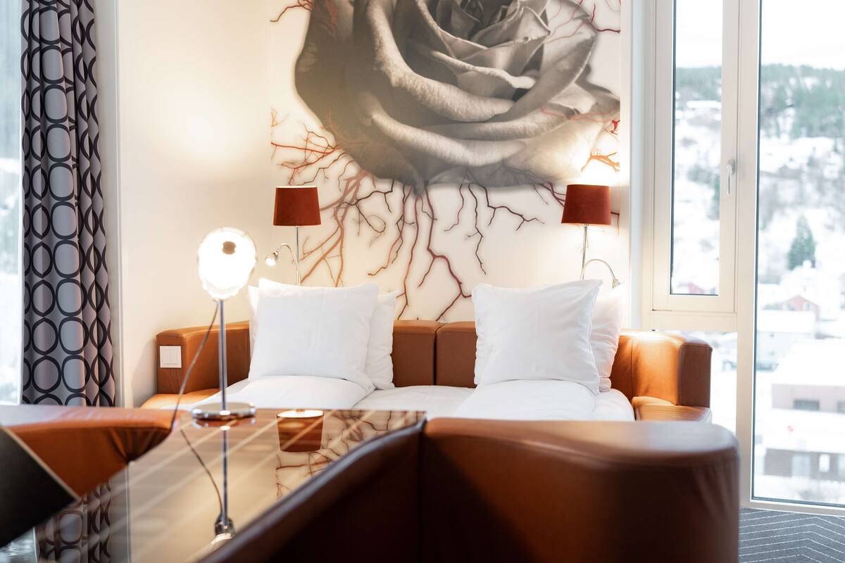 A hotel room with a bed, a nightstand, a lamp, and a large window with a view of buildings.
