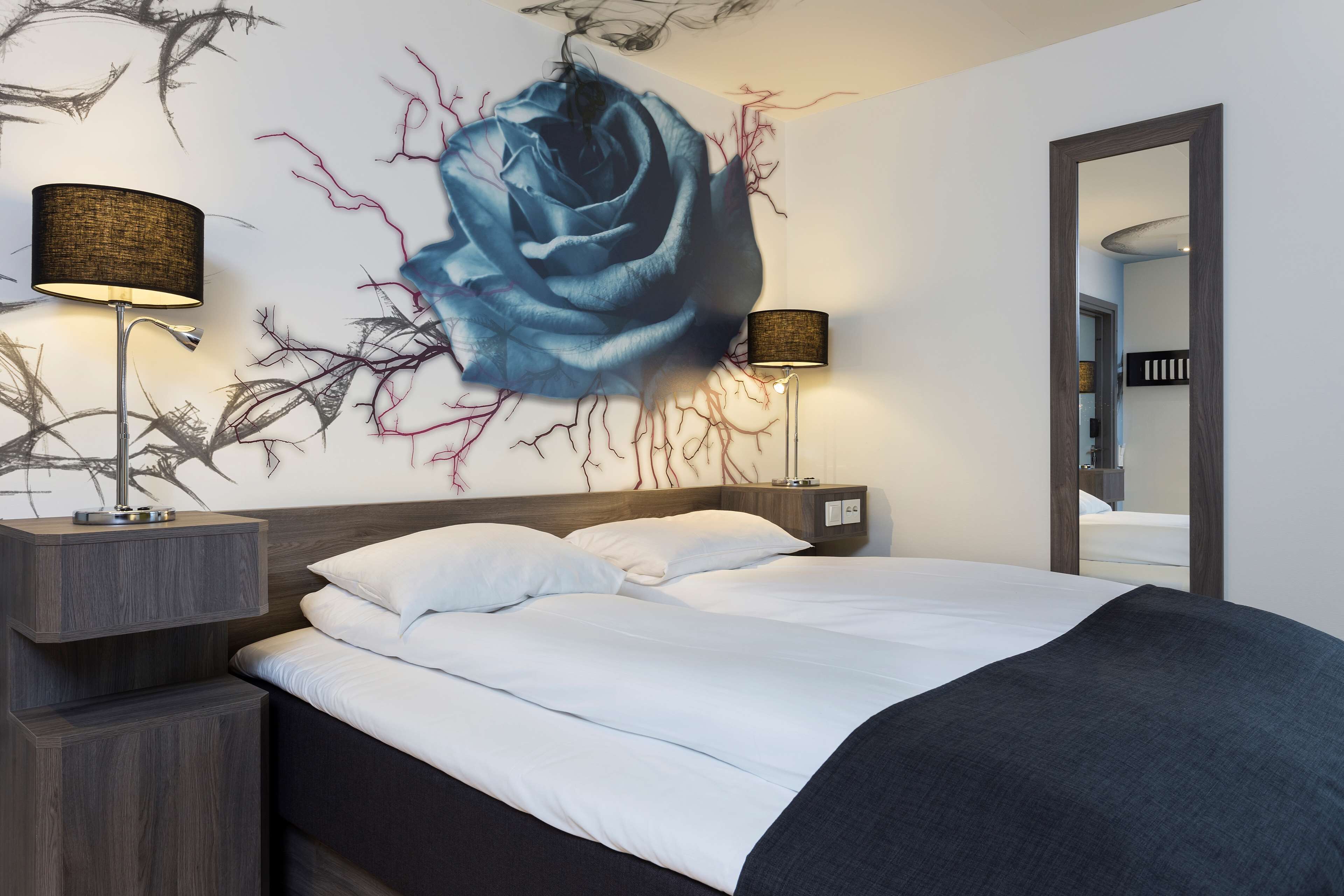 A modern hotel room with a large bed, two bedside tables with lamps, a wall mural of a blue rose, and a mirror on the wall.