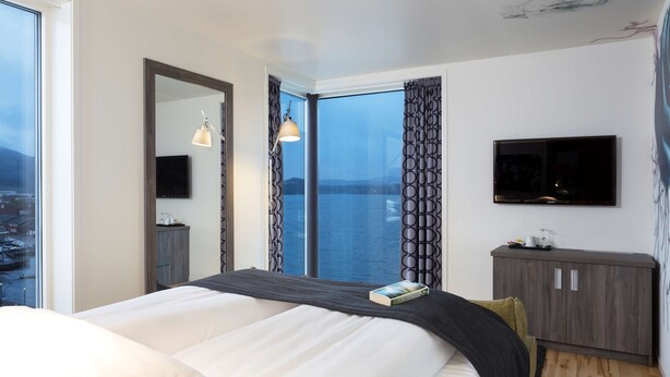 A modern hotel room with a large bed, a TV mounted on the wall, and a view of the water. - Scandic Rock City (Namsos)