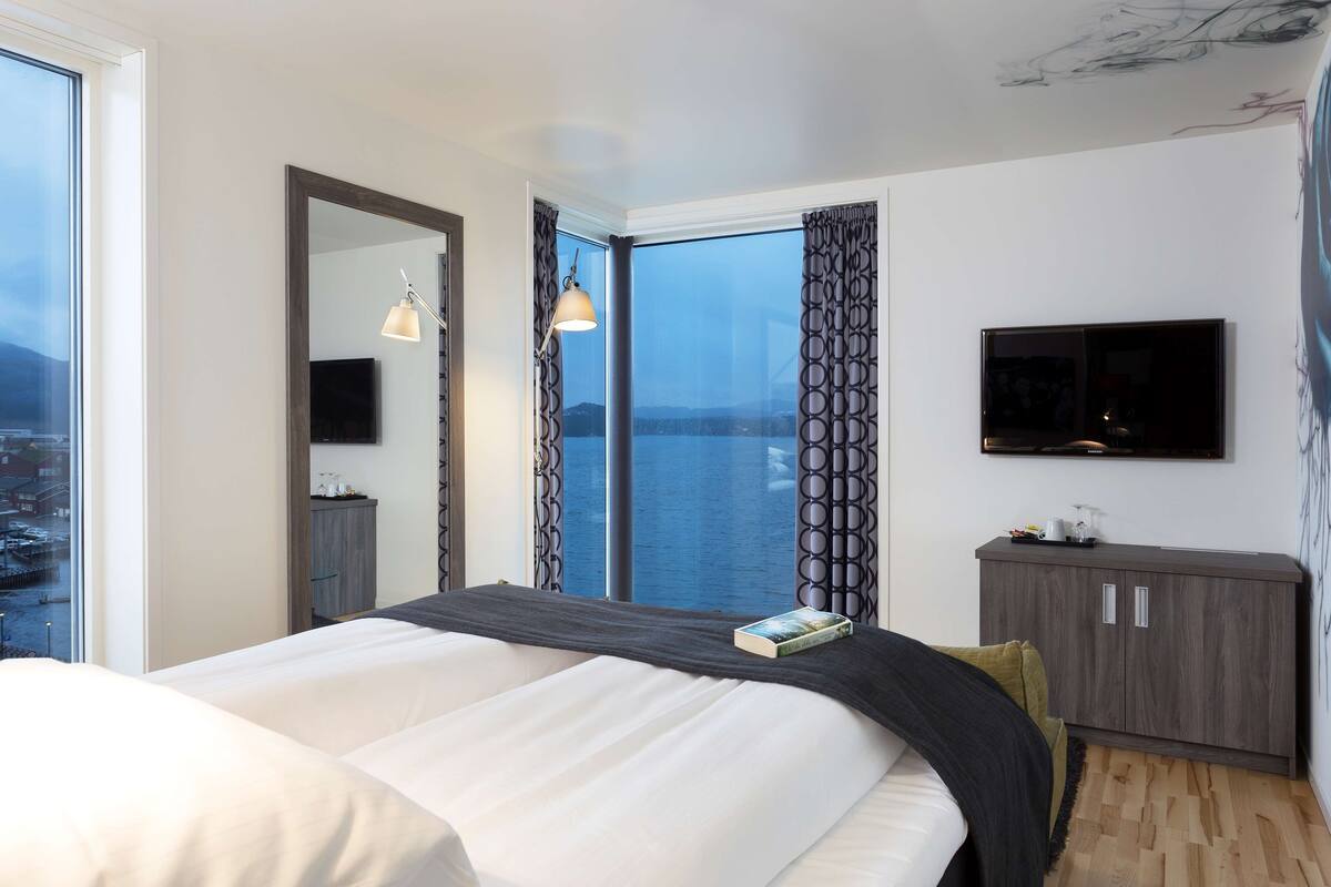 A modern hotel room with a large bed, a TV mounted on the wall, and a view of the water.