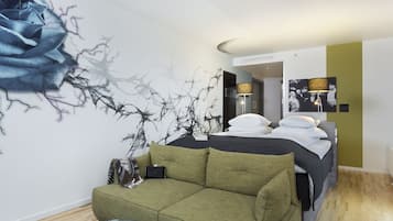 A modern bedroom with a green sofa, a bed with white bedding, a large abstract wall art, and a lamp.
