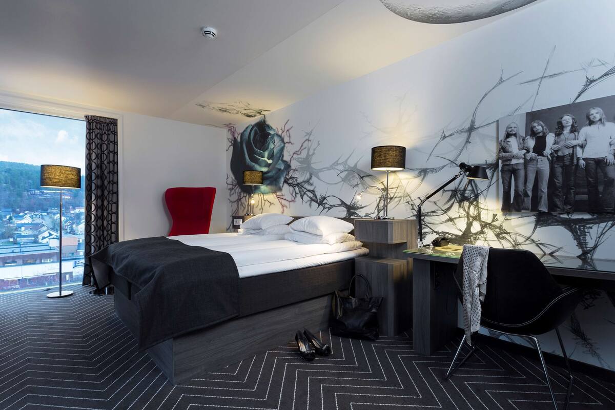 A modern hotel room with a large bed, a desk with a chair, a red armchair, and a city view through the window.