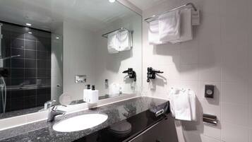 Un baño moderno con un espejo grande, azulejos blancos, encimera de granito negro, lavabo blanco, inodoro negro y zona de ducha con mampara de cristal.