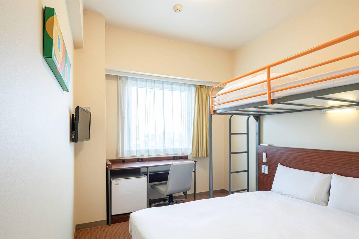 A hotel room with a bunk bed, a desk with a chair, a small refrigerator, and a window with curtains.