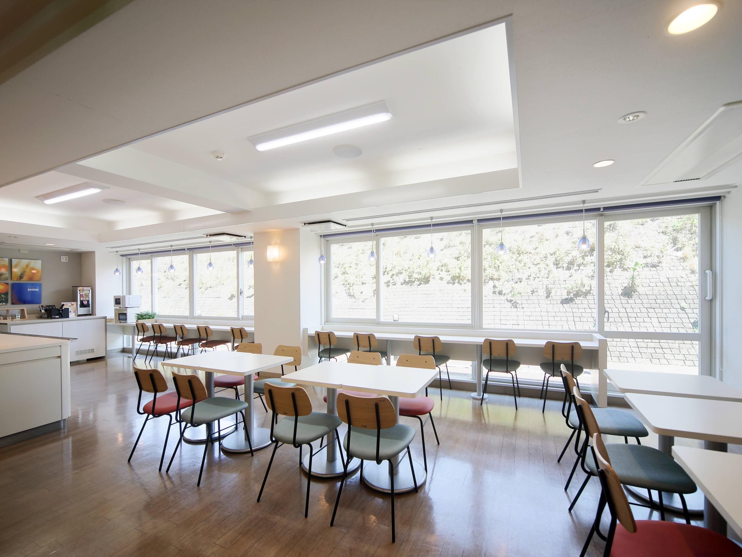 Un vaste espace dédié à la salle à manger, avec de nombreuses tables et chaises, de grandes fenêtres laissant entrer la lumière naturelle et une cuisine visible au fond.