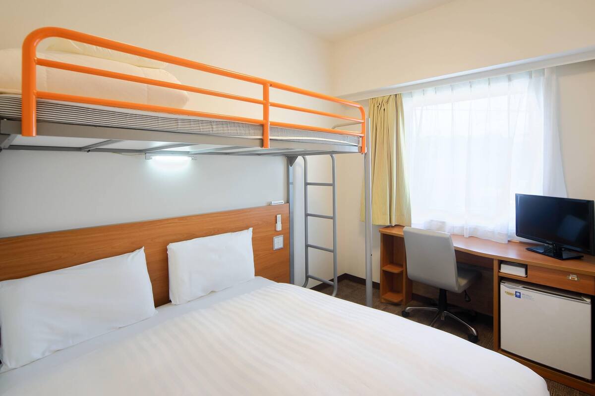 A hotel room with a bunk bed, a desk with a computer, a television, and a mini-fridge.