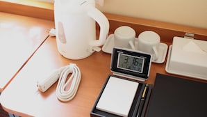 Una mesa con un hervidor eléctrico blanco, dos tazas blancas, un reloj digital, un cable de alimentación blanco, una libreta negra y un bolígrafo.