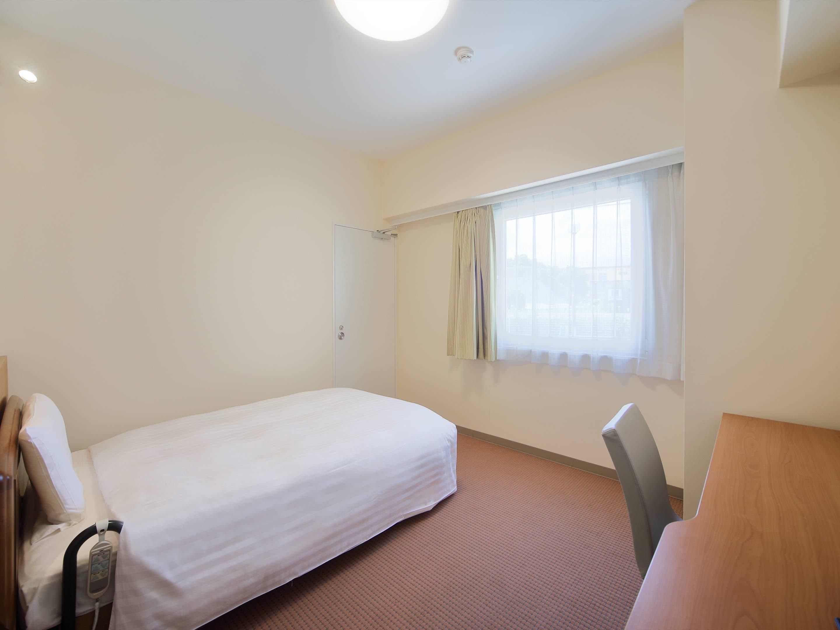 A hotel room with a single bed, a bedside table, a chair, a window with curtains, and a door.