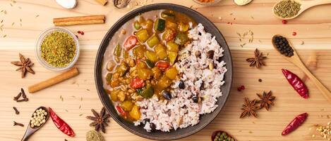 A bowl of rice and vegetable curry surrounded by various spices and ingredients on a wooden surface.