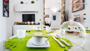 Una cocina moderna con una mesa de comedor preparada para dos personas, que incluye una tetera blanca, tazas y una campana de cristal cubriendo un pastel.