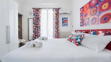 Quarto de hotel moderno com uma cama grande, cortinas florais, cabeceira vermelha e obras de arte coloridas nas paredes.