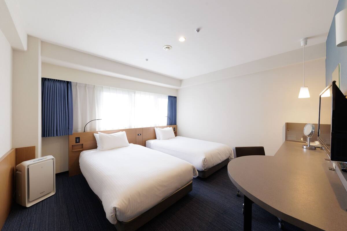 A hotel room with two beds, a desk, a chair, a small table, a mirror, and a window with curtains.