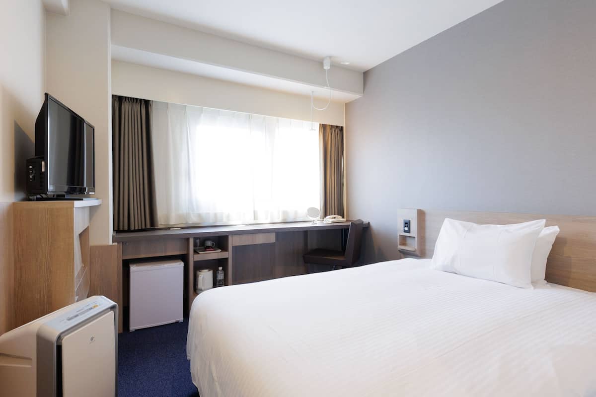 A hotel room with a bed, a desk, a television, a mini-fridge, and a window with curtains.