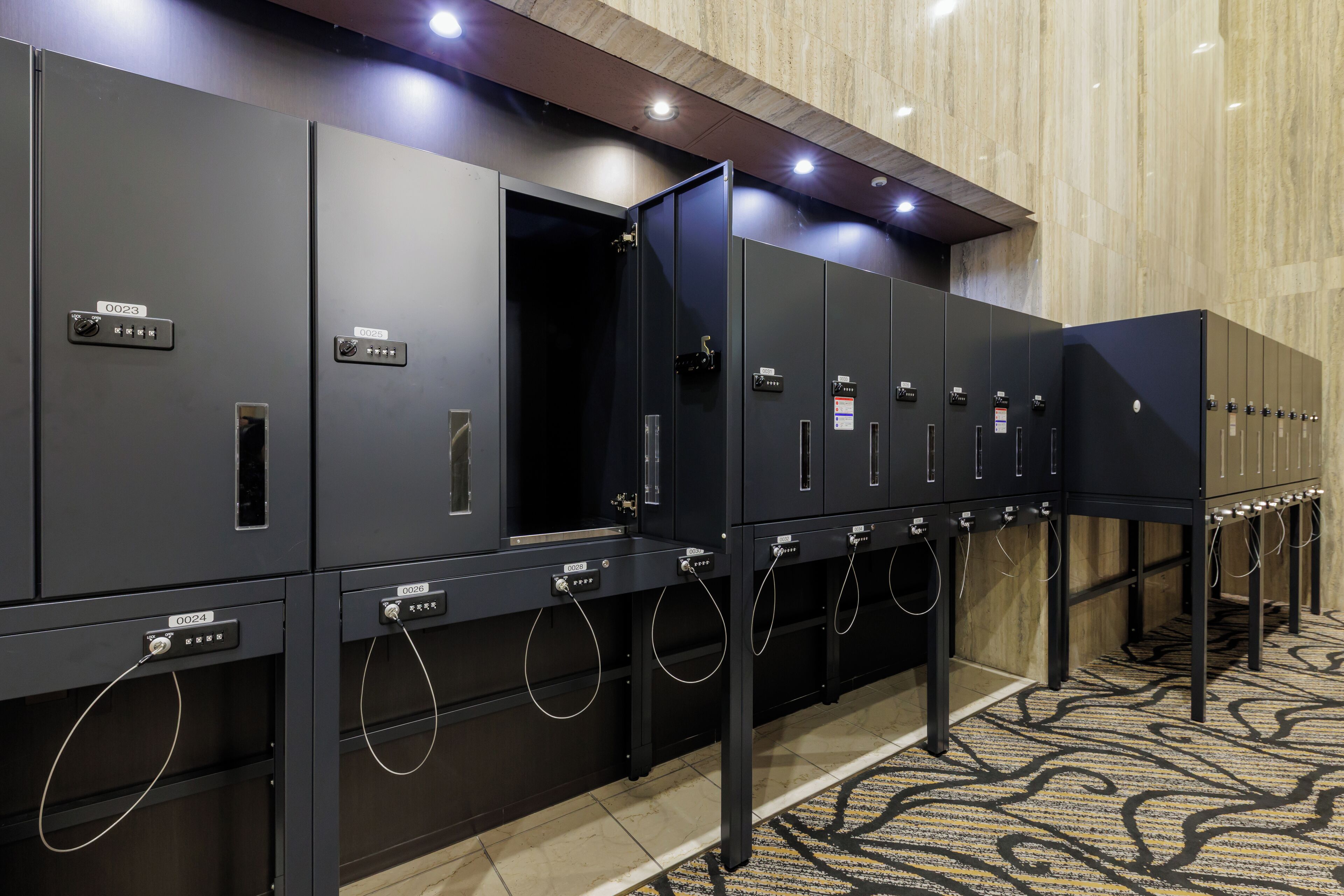 A row of black lockers with numbered labels, a carpeted floor, and a marble wall in the background.
