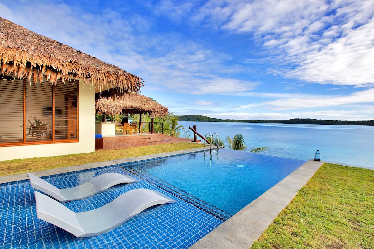Una piscina con sillas de descanso, una estructura con techo de paja y vista al agua.