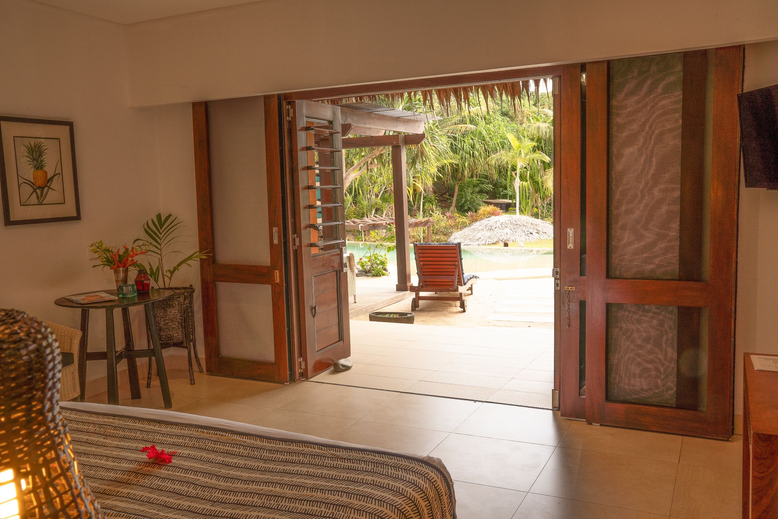 A room with a bed, a small table with a plant, and a door leading to an outdoor area with a chair and a thatched structure.