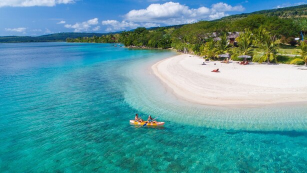 Una playa con agua turquesa cristalina, una orilla arenosa y algunas personas en kayak.