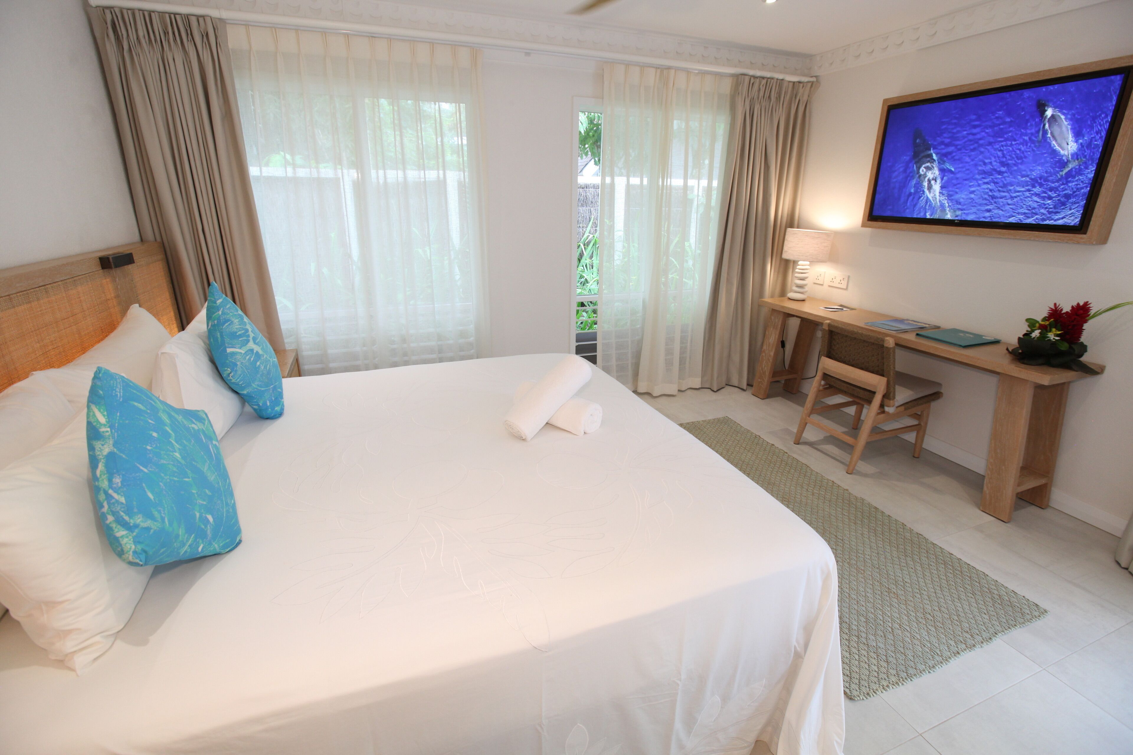 A hotel room with a large bed, a desk, a television, and a view of greenery outside.