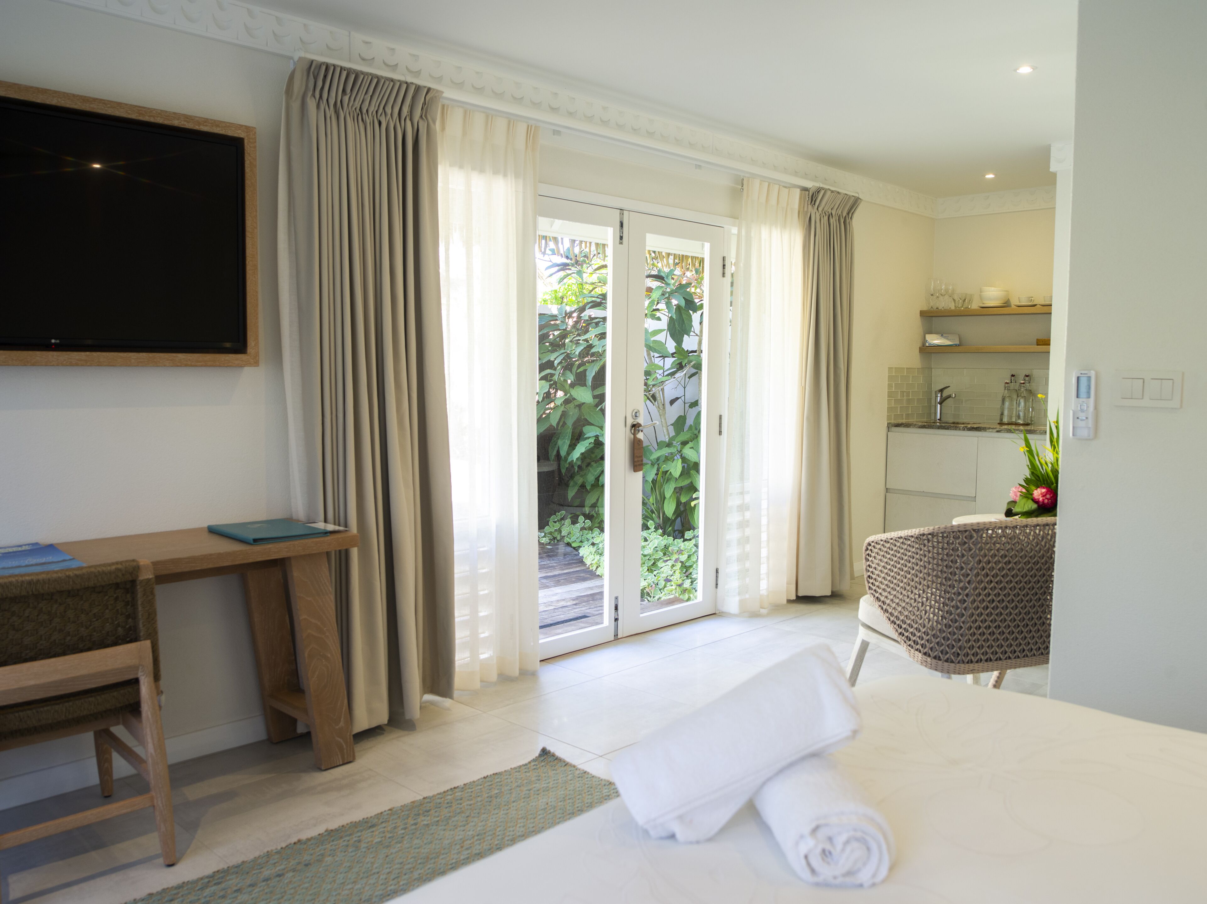 Habitación de hotel con cama, escritorio, silla, televisor y puerta corrediza de cristal que da a un jardín.