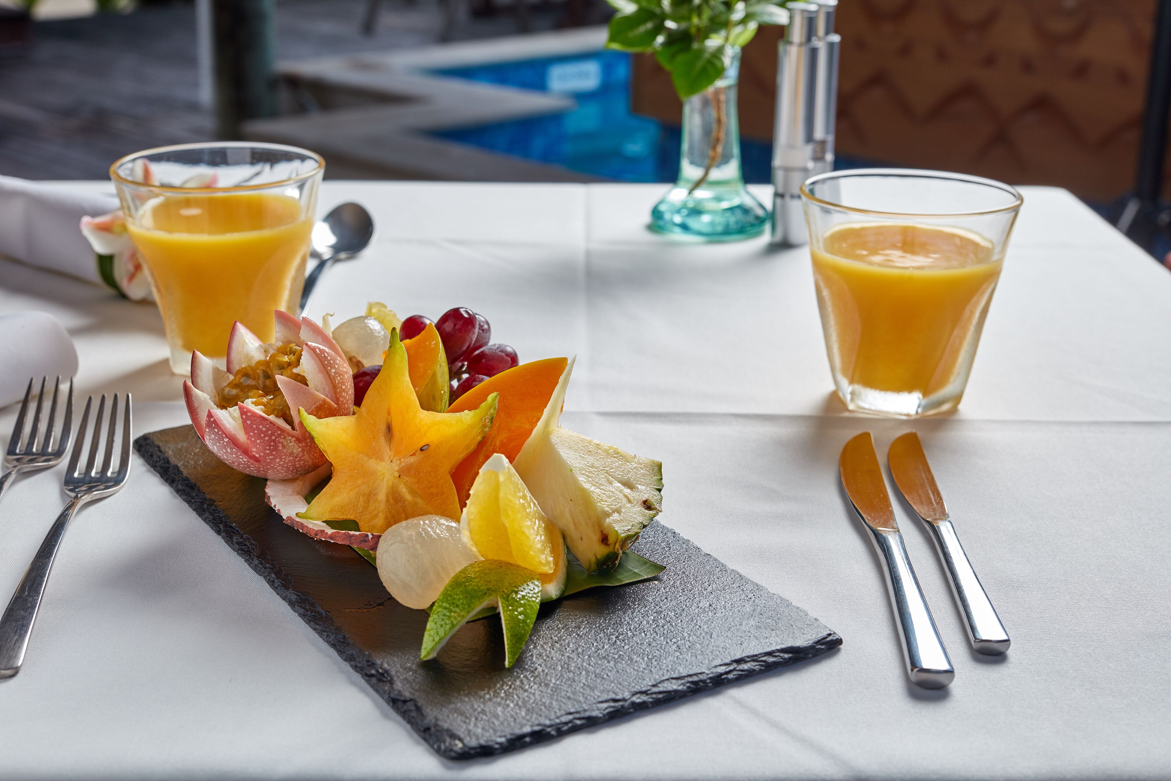 Ontbijttafel met fruit, sap op een arduinen bord, bestek en uitzicht op het zwembad.