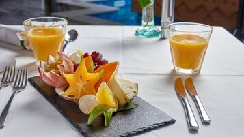 Ontbijttafel met fruit, sap op een arduinen bord, bestek en uitzicht op het zwembad.