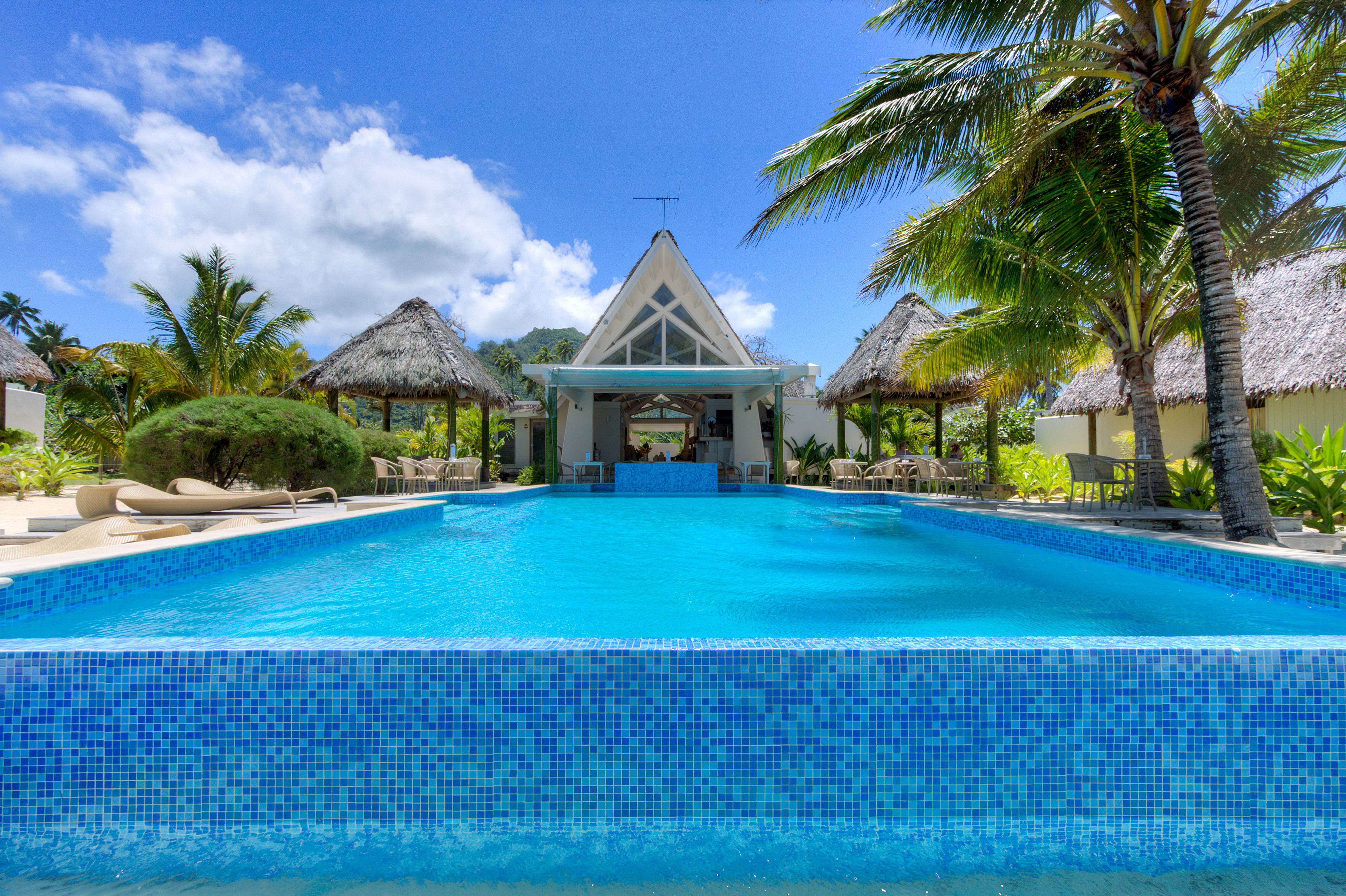 A swimming pool with a clear blue tile border, surrounded by palm trees and thatched-roof structures.