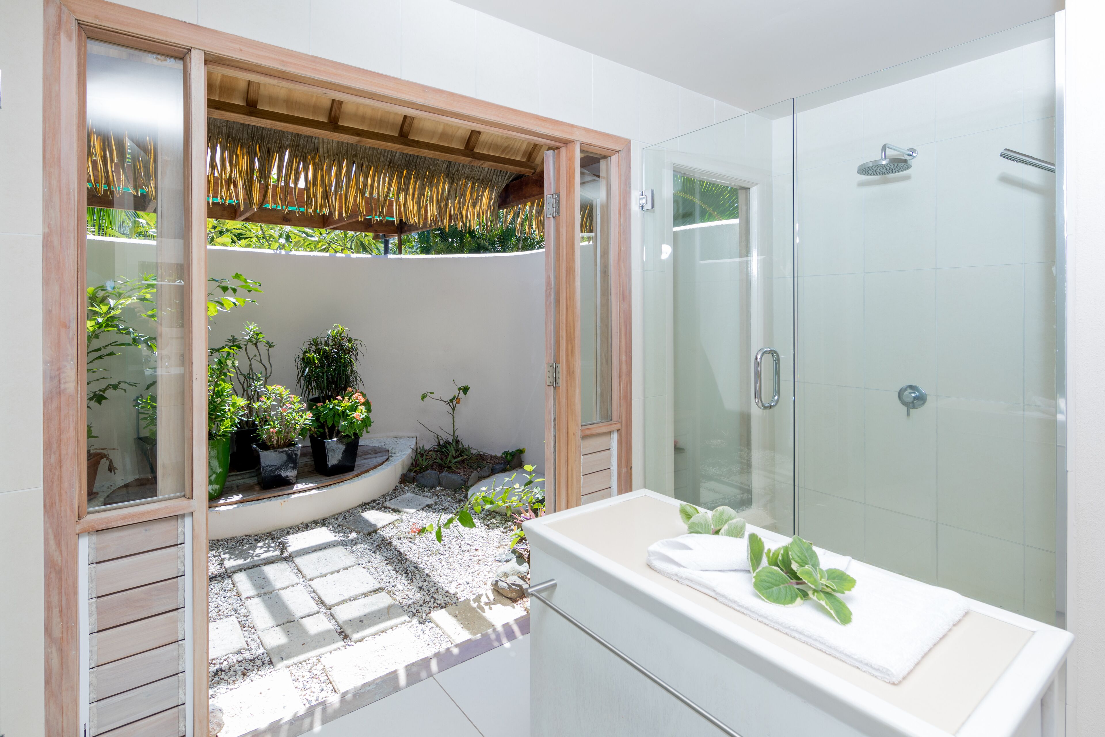 Une salle de bain moderne avec une douche à l’italienne en verre, une baignoire blanche et un petit espace jardin visible à travers une fenêtre à cadre en bois.