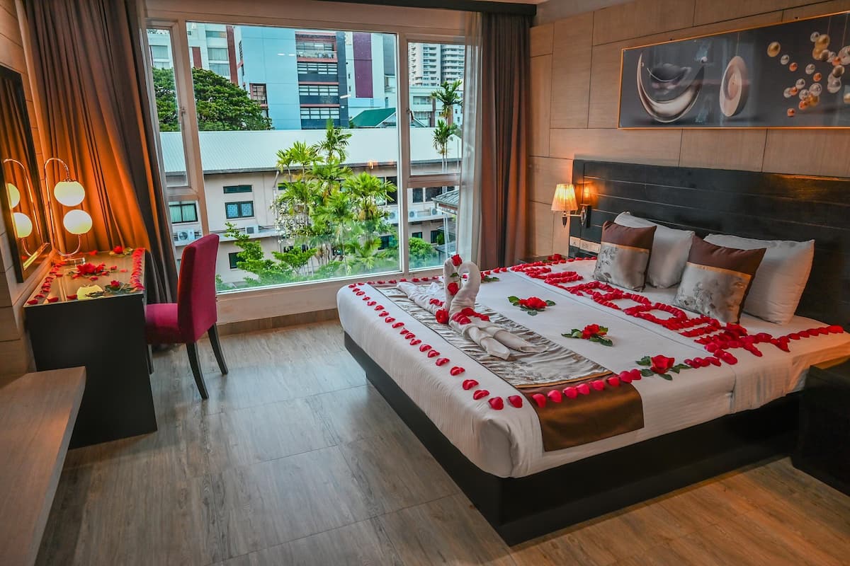 A hotel room with a large bed adorned with rose petals, a red chair, a vanity with a mirror, and a view of buildings and greenery outside.