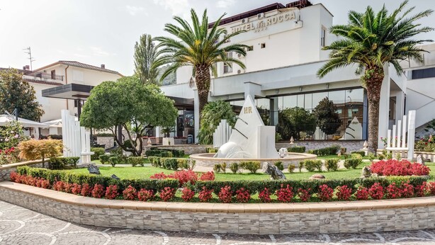 A modern hotel with a landscaped garden, palm trees, and a fountain.
