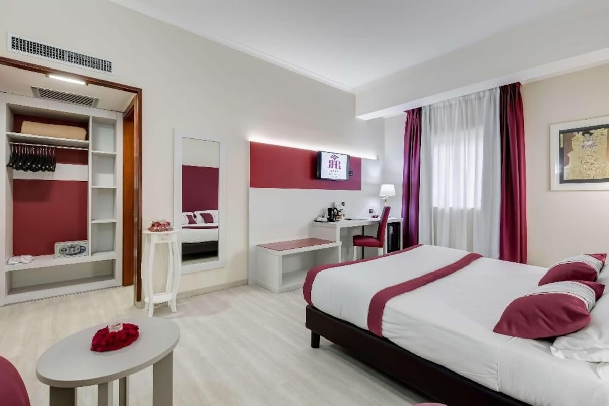 A hotel room with a bed, a desk, a chair, a wardrobe, and a small table.