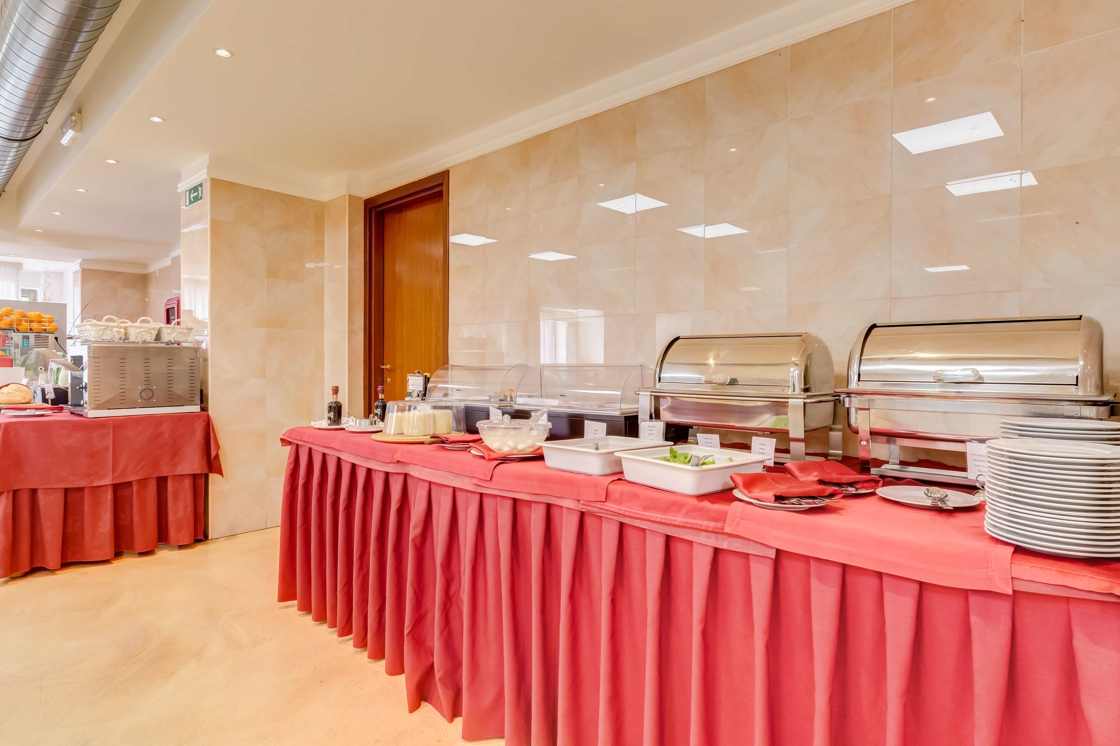 Ein reichhaltiges Buffet mit verschiedenen Speisen und Besteck auf einer roten Tischdecke, in einem gut beleuchteten Raum mit gefliesten Wänden und einer Holztür.