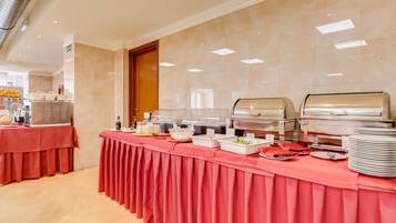 Ein reichhaltiges Buffet mit verschiedenen Speisen und Besteck auf einer roten Tischdecke, in einem gut beleuchteten Raum mit gefliesten Wänden und einer Holztür.