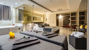 A modern hotel room with a large bed, two smaller beds, a sitting area, a dining area, and a bathroom with a large mirror.