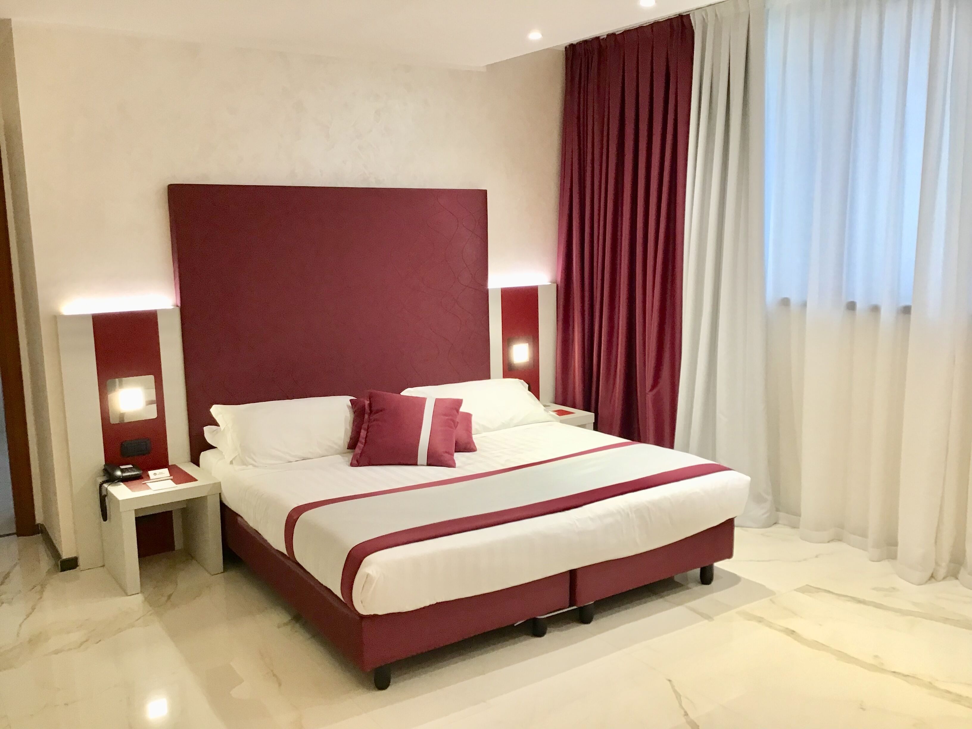 A hotel room with a large bed, a nightstand, a red headboard, and a window with curtains.