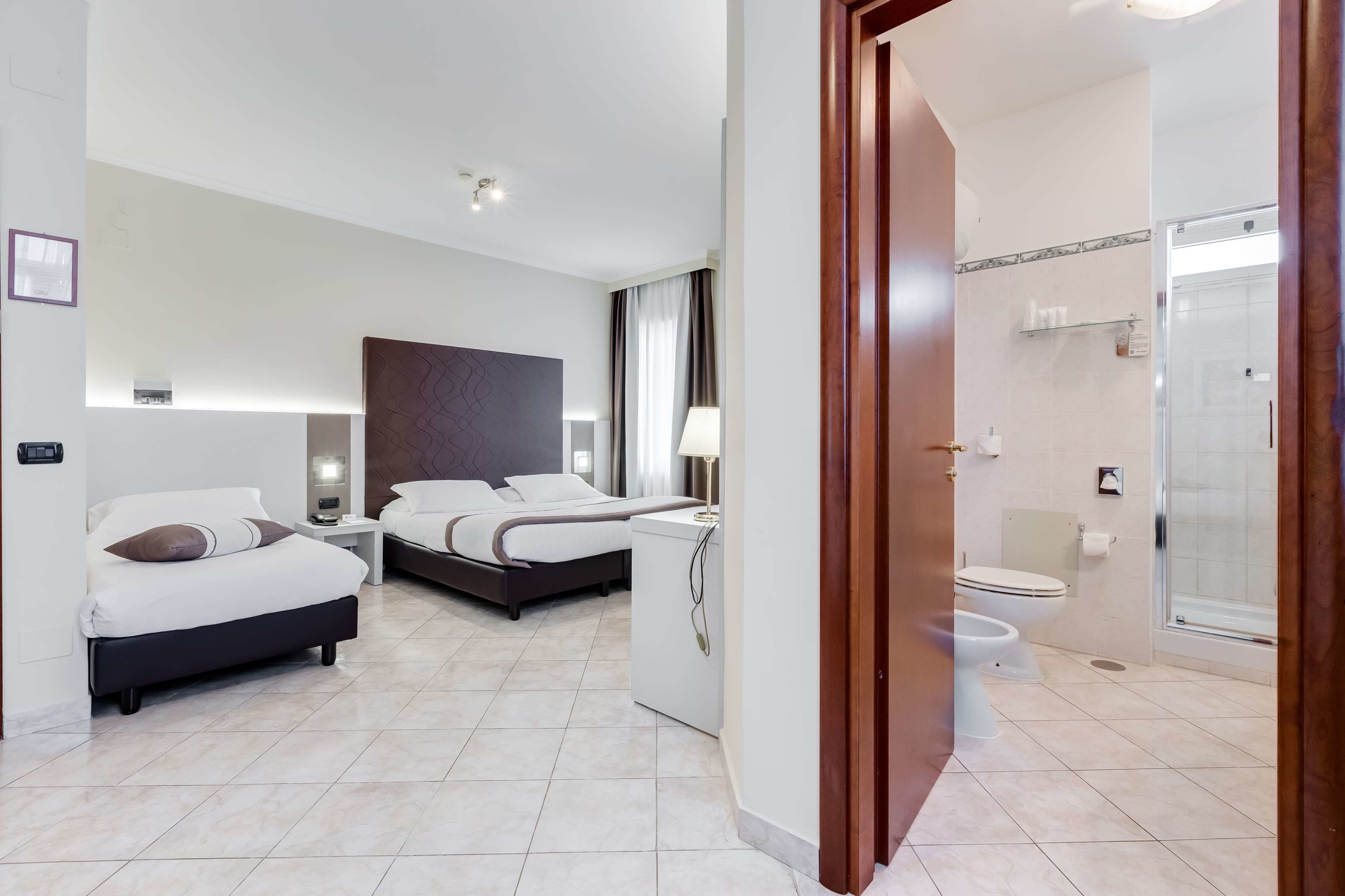 A hotel room with two beds, a bedside table, a lamp, a shower area, and a toilet.