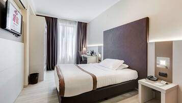 Une chambre d’hôtel moderne avec un grand lit, une table de chevet avec un téléphone et des documents, un bureau avec une lampe et un téléviseur fixé au mur.