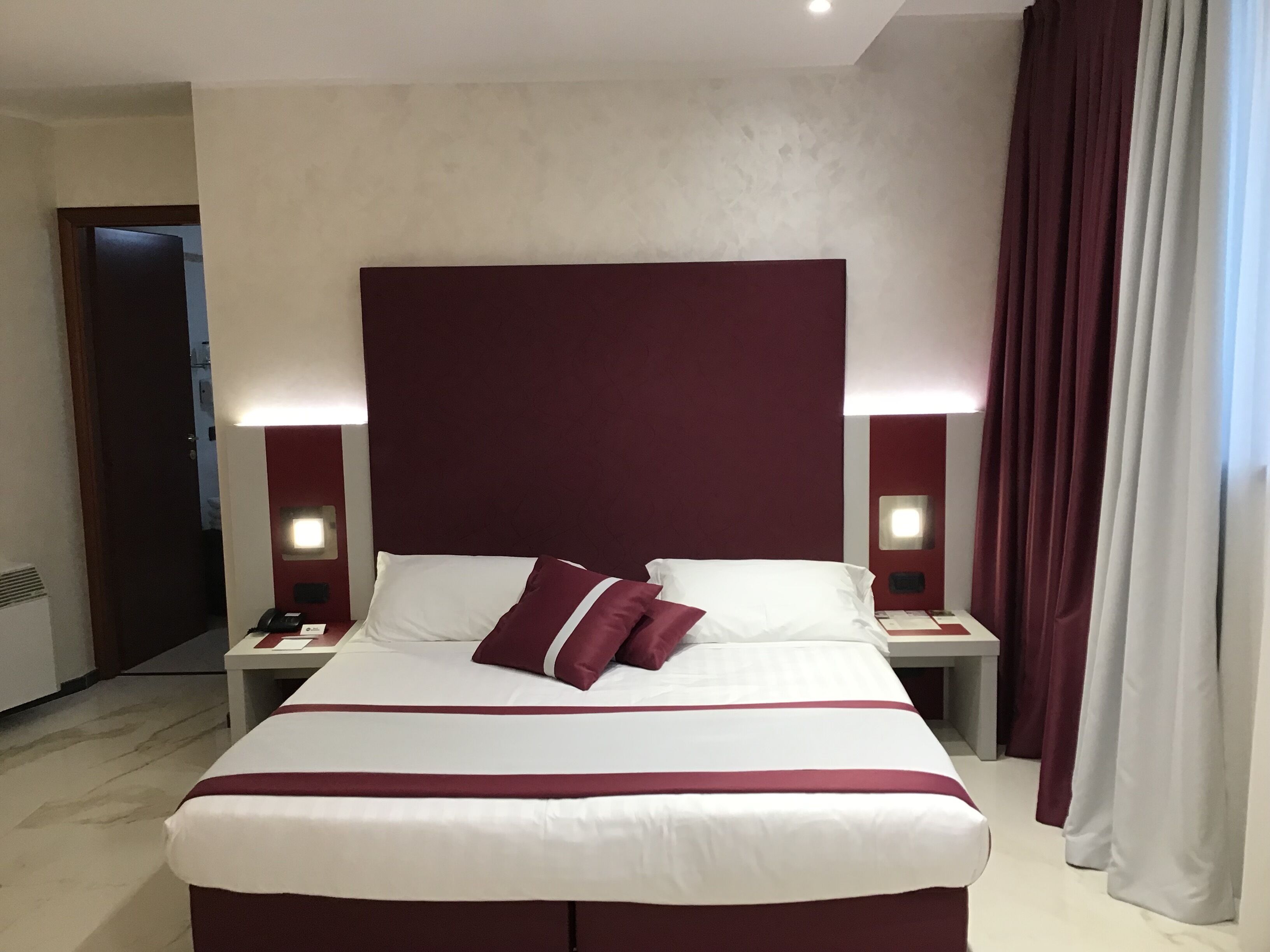 Ein Hotelzimmer mit einem großen Bett, zwei Nachttischen, einem Schminktisch und einem roten Kopfteil sowie einem Fenster mit Vorhängen.