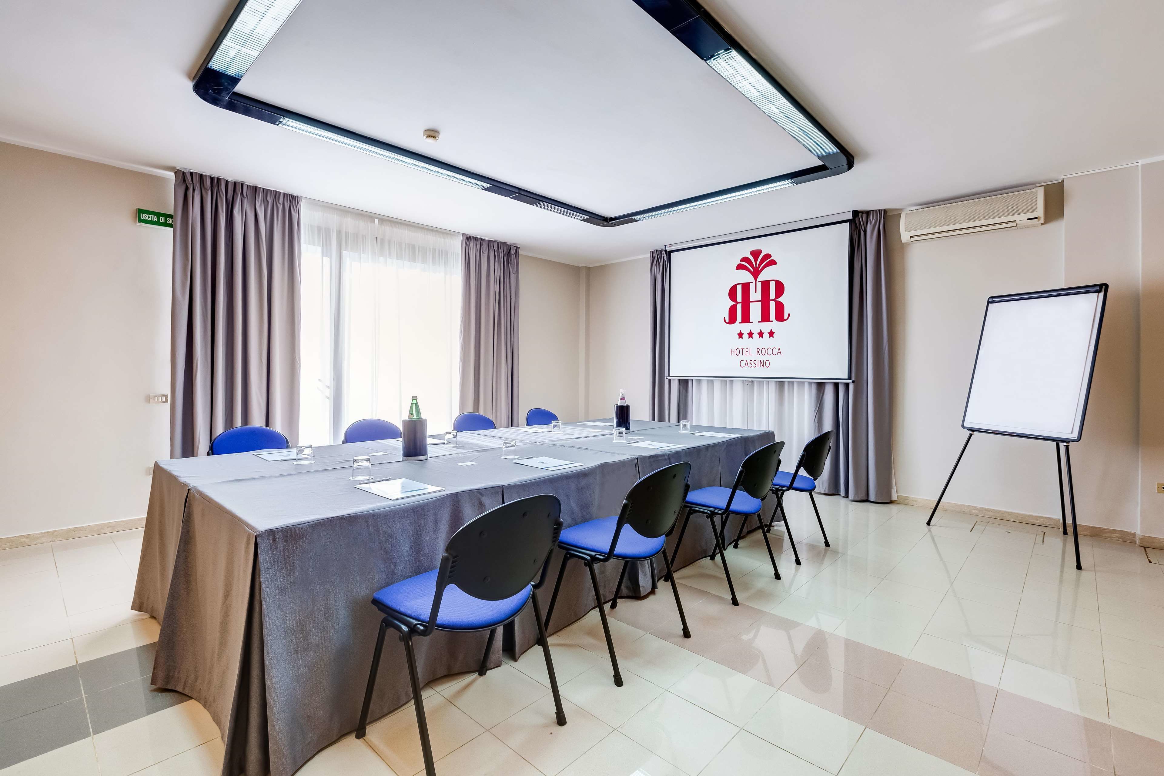 Ein Konferenzraum mit langem Tisch, blauen Stühlen, einer Whiteboard-Tafel und einer Projektionsleinwand, auf der das Hotel-Logo zu sehen ist.