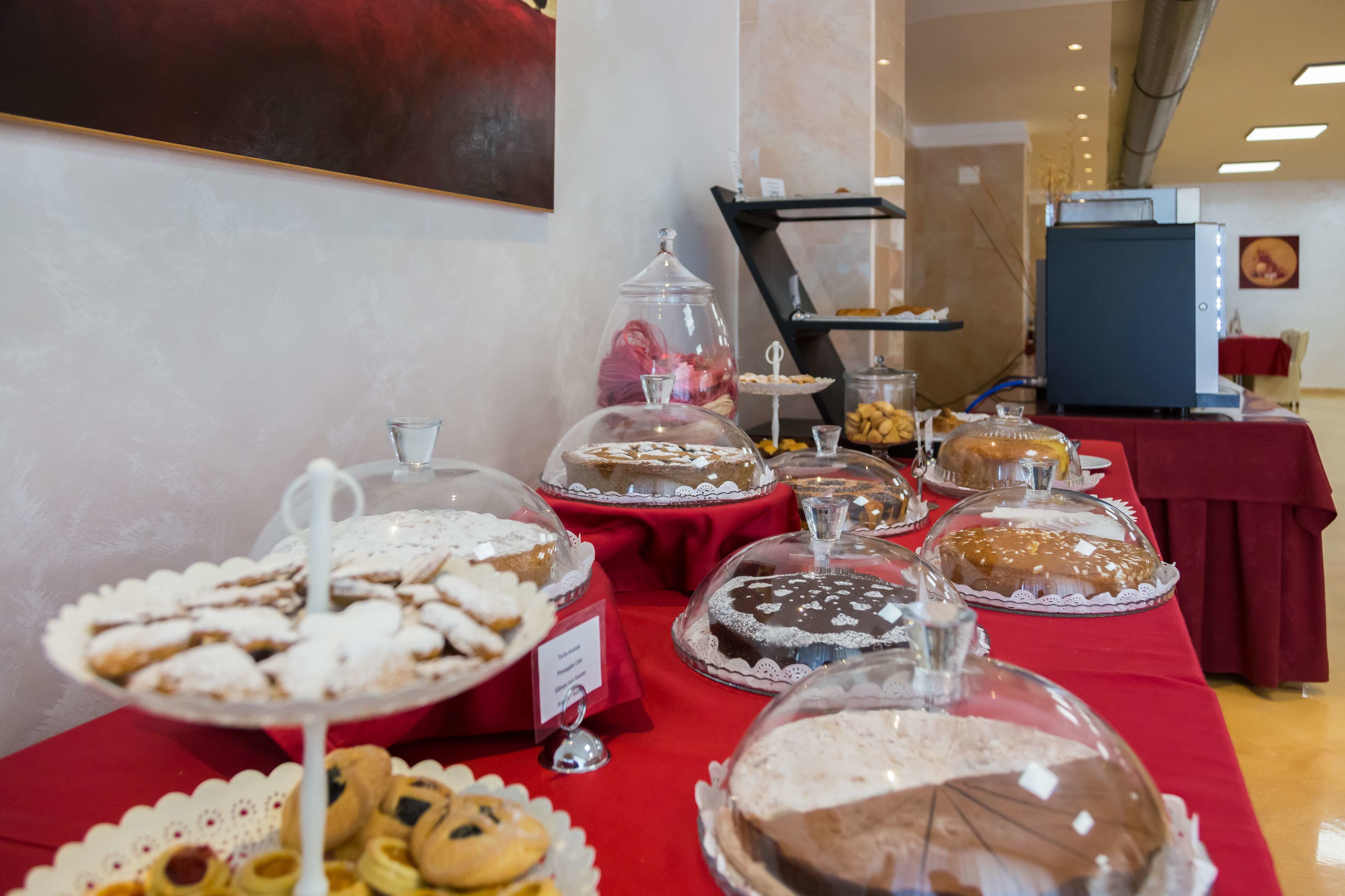 A buffet table with various desserts covered in glass domes, a red tablecloth, and a sign indicating a dessert station.