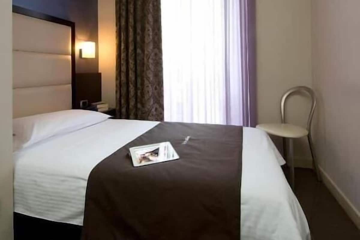 A hotel room with a bed, a nightstand, a chair, and a window with curtains.