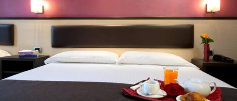 Habitación de hotel con cama, mesitas de noche, una bandeja con alimentos para el desayuno y un tocador con teléfono y libros.