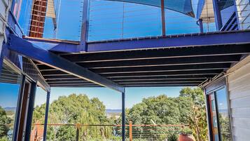 A covered outdoor seating area with a table and chairs, a blue shade sail, and a view of trees and hills.