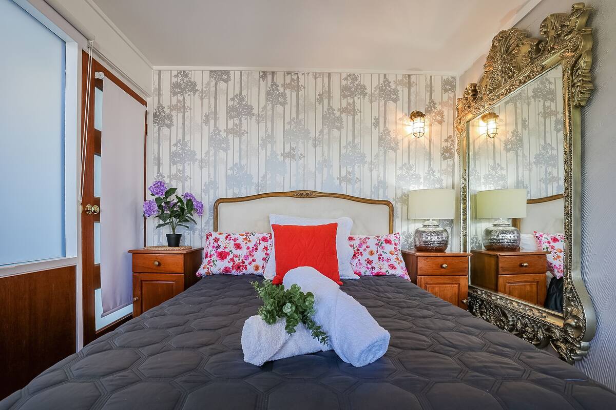A neatly made bed with a dark quilt, a red pillow, and a white towel with greenery. There are two bedside tables with lamps, a large ornate mirror, and a wall with vertical stripes.