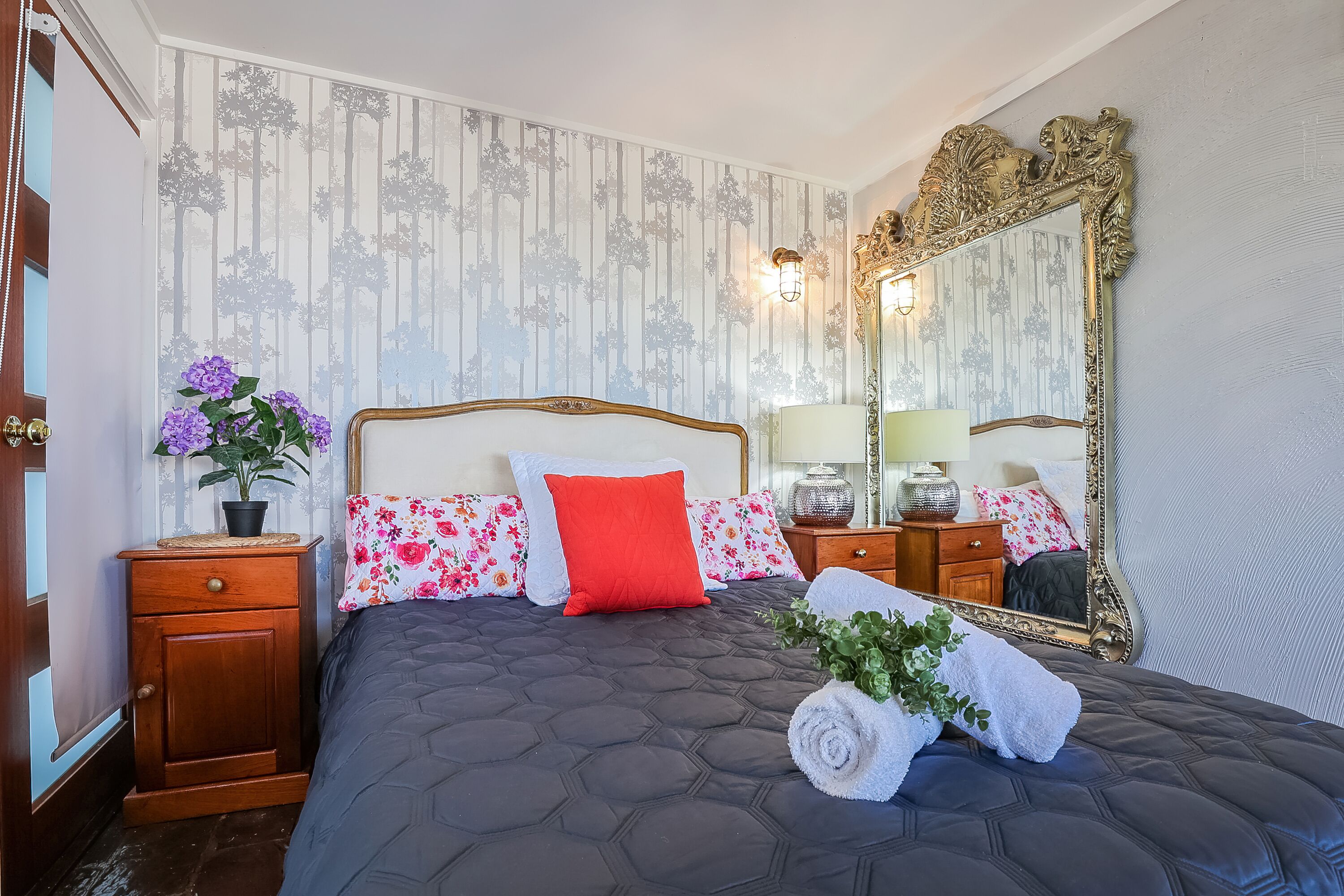 A neatly arranged bedroom with a large bed, floral-patterned pillows, a wooden nightstand, a mirror, and a potted plant.